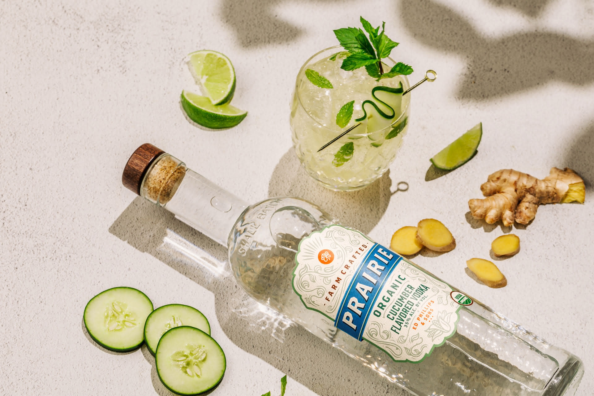 Prairie Organic Cucumber Vodka bottle surrounded by cucumber, lime, mint, and ginger with a cocktail